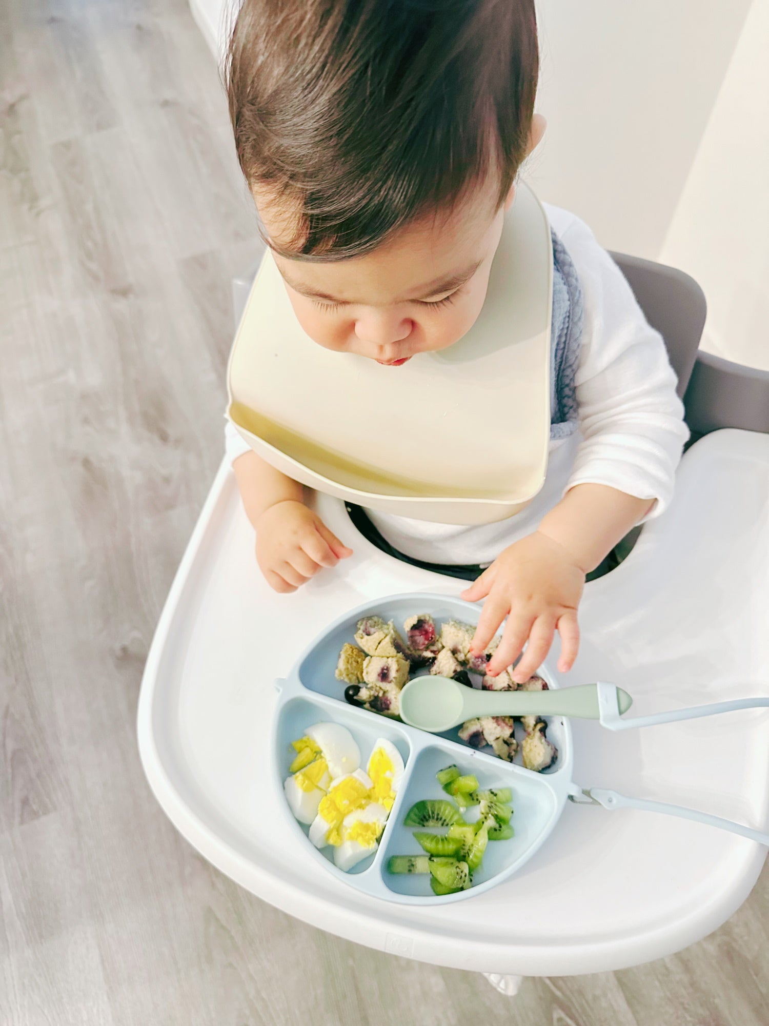Child in a high chair with a dropless divided plate and utensils, eating a meal.