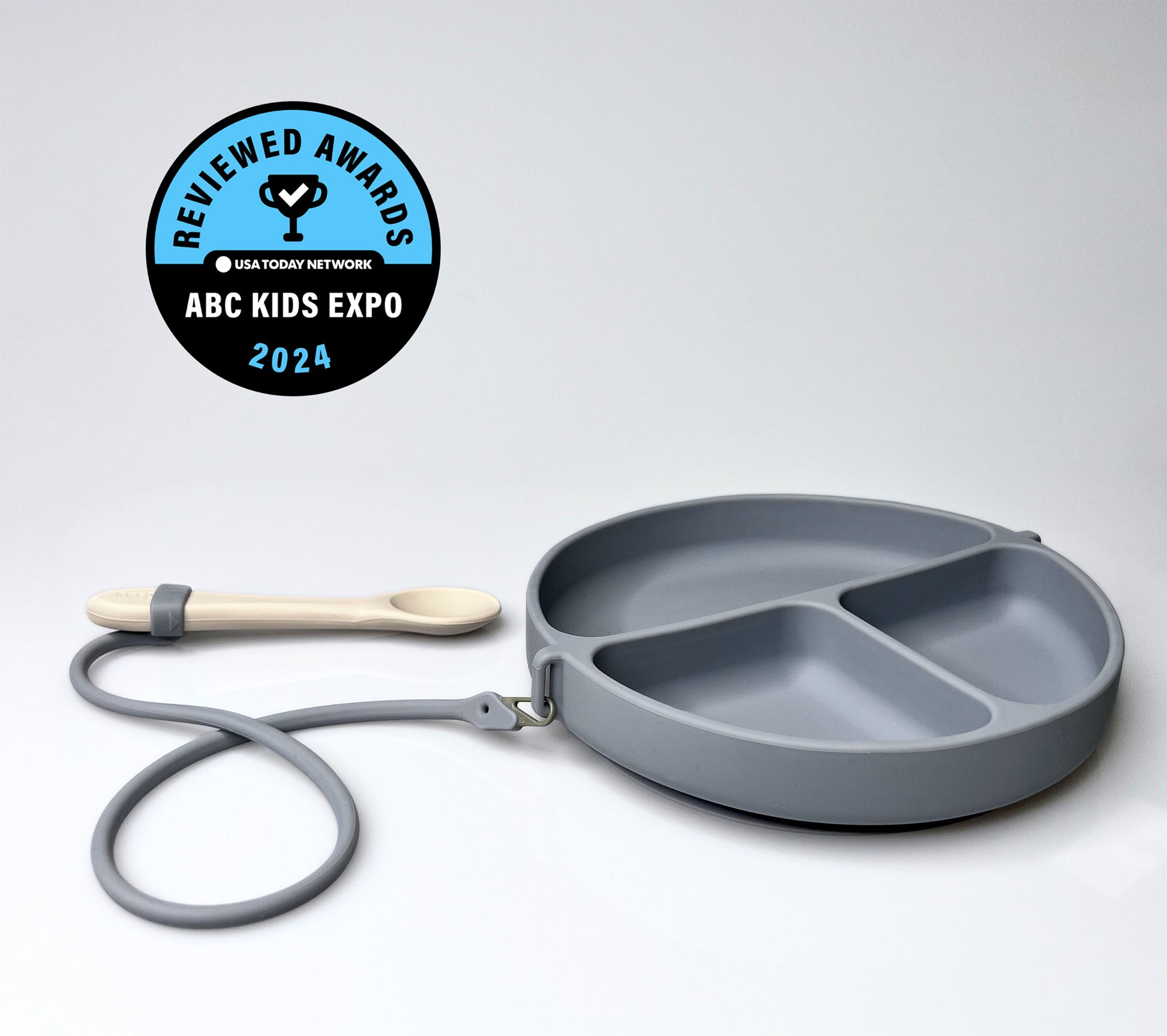 Gray children's plate with divided sections and a silicone spoon on a white background, featuring the ABC Kids Expo 2024 badge.