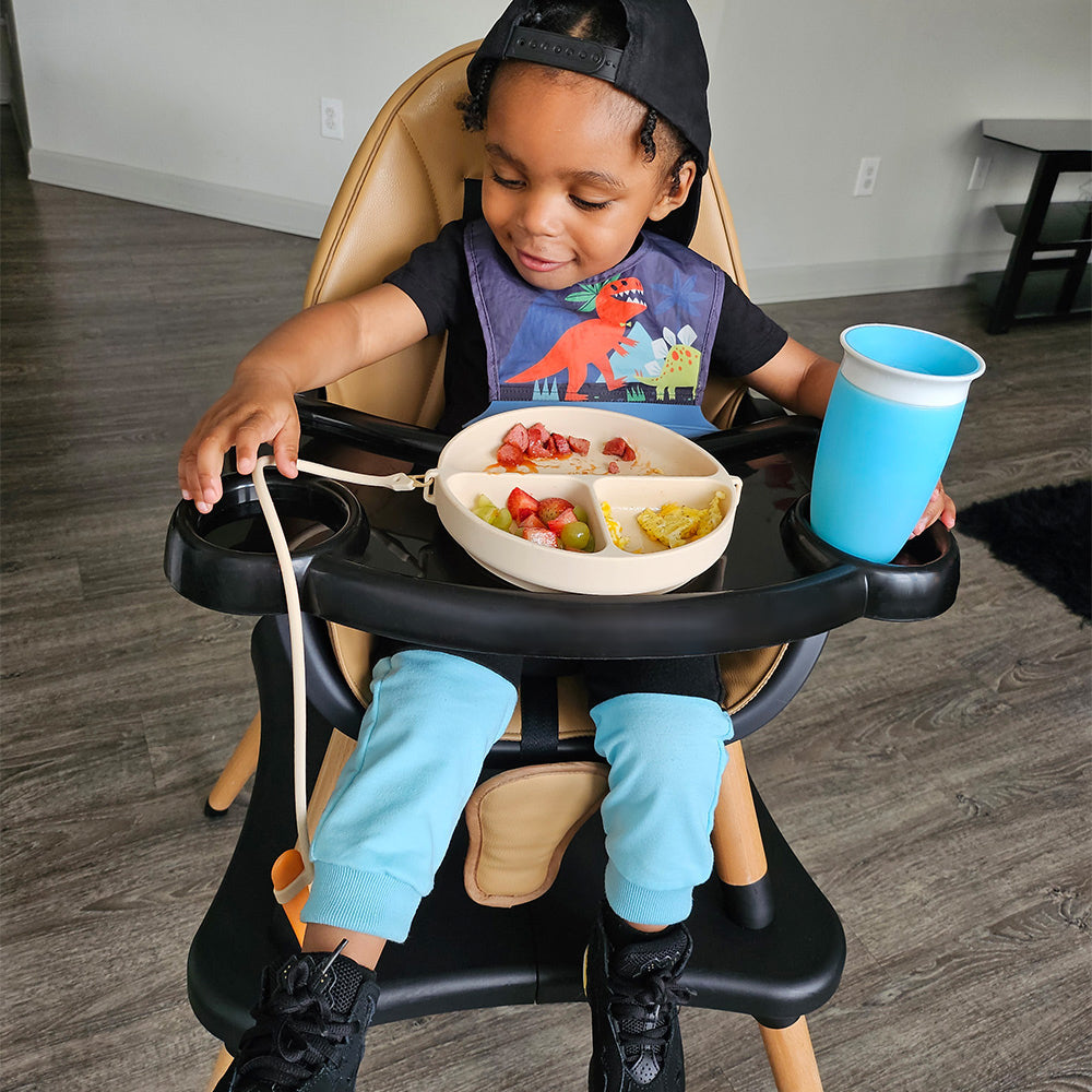 An african american toddler boy using dropless plate on a highchair dropping his fork with a drinking cup