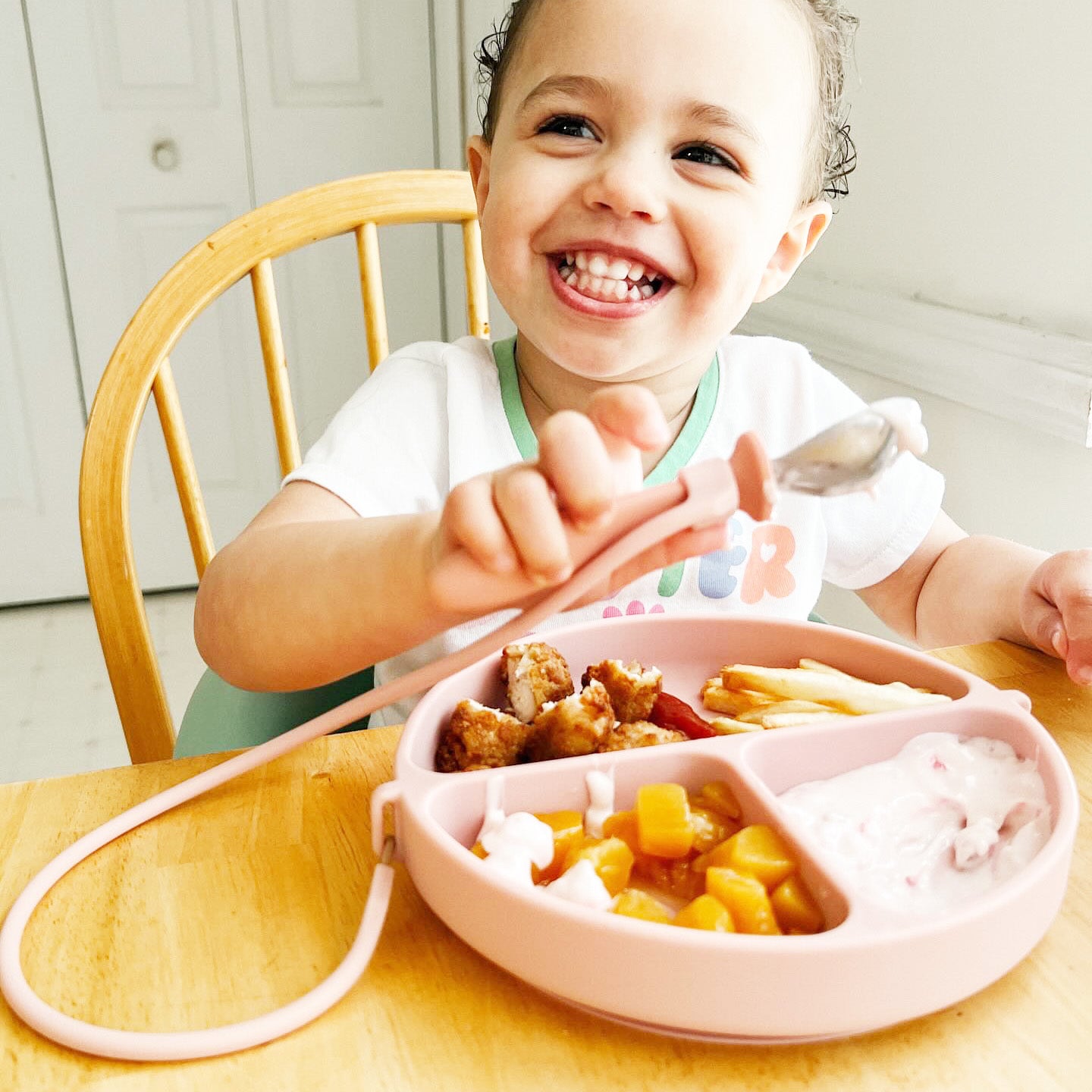 Toddler girl using a pink dropless plate on a wooden table with a utensil being attached to the handle on the plate